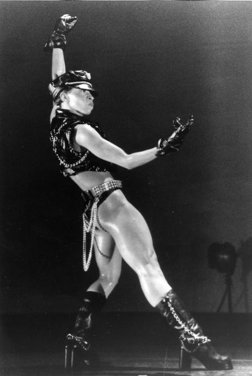 A woman in a biker-inspired costumed (leather hat, gloves, vest, briefs and boots) strikes a powerful pose. One arm is extended above her head in a fist and the other is gesturing in front of her.